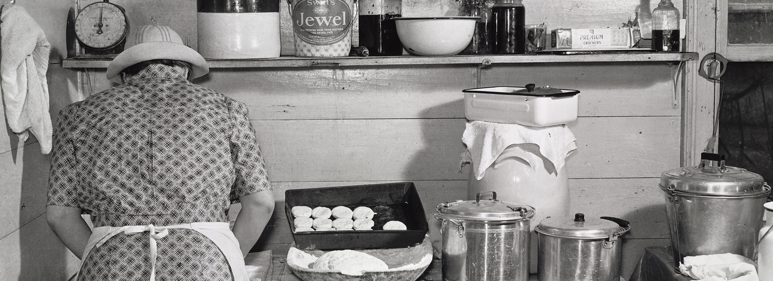 A black and white photo of a kitchen with pots, pans, and other items with a person in a print dress, hat, and apron seen from behind