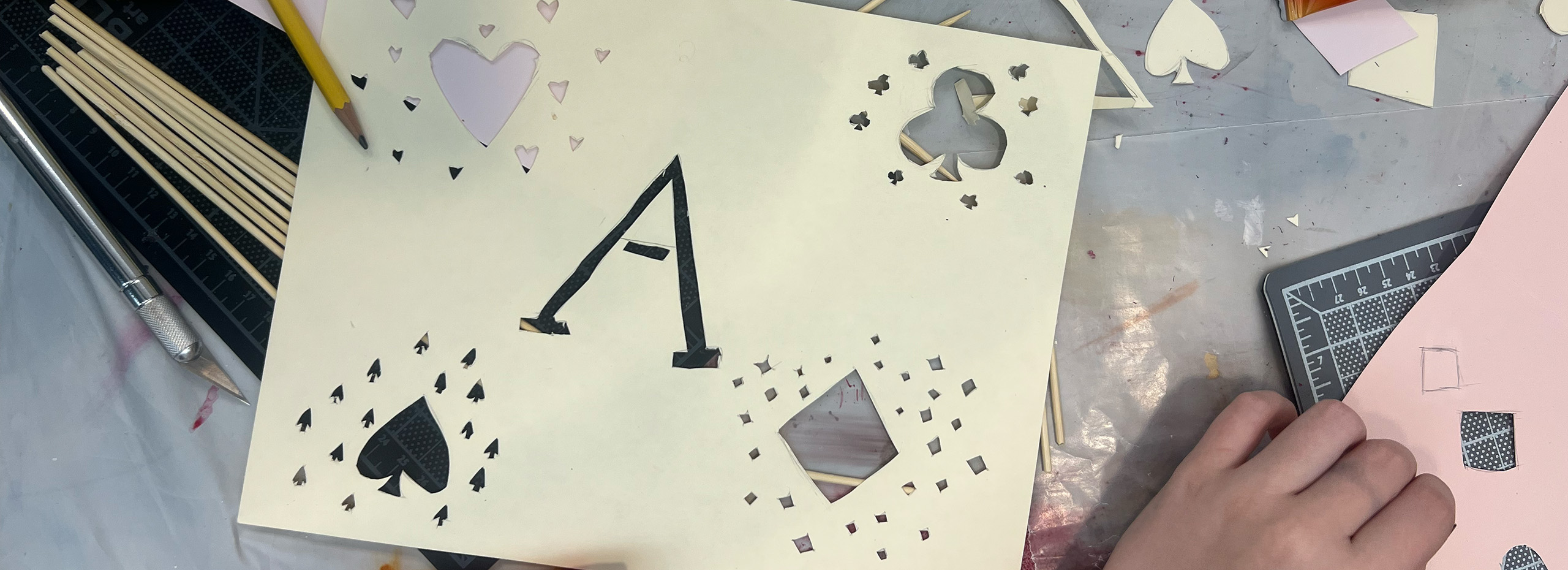 A craft table with paper cutouts, pencils, and a cutting tool