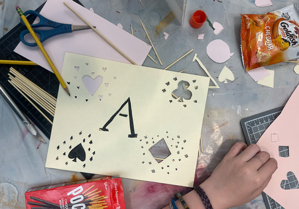 A craft table with a hand, snacks, paper cutouts, pencils, and a cutting tool