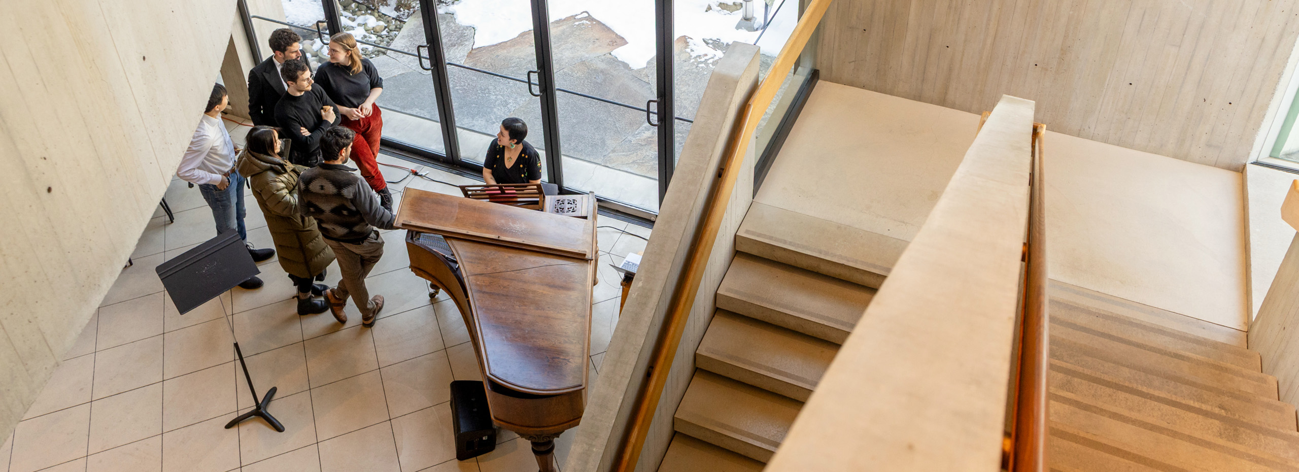 People gather around a piano next to a concrete staircase and glass doors facing outside