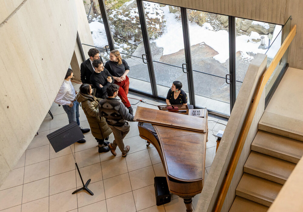 People gather around a piano next to a concrete staircase and glass doors facing outside