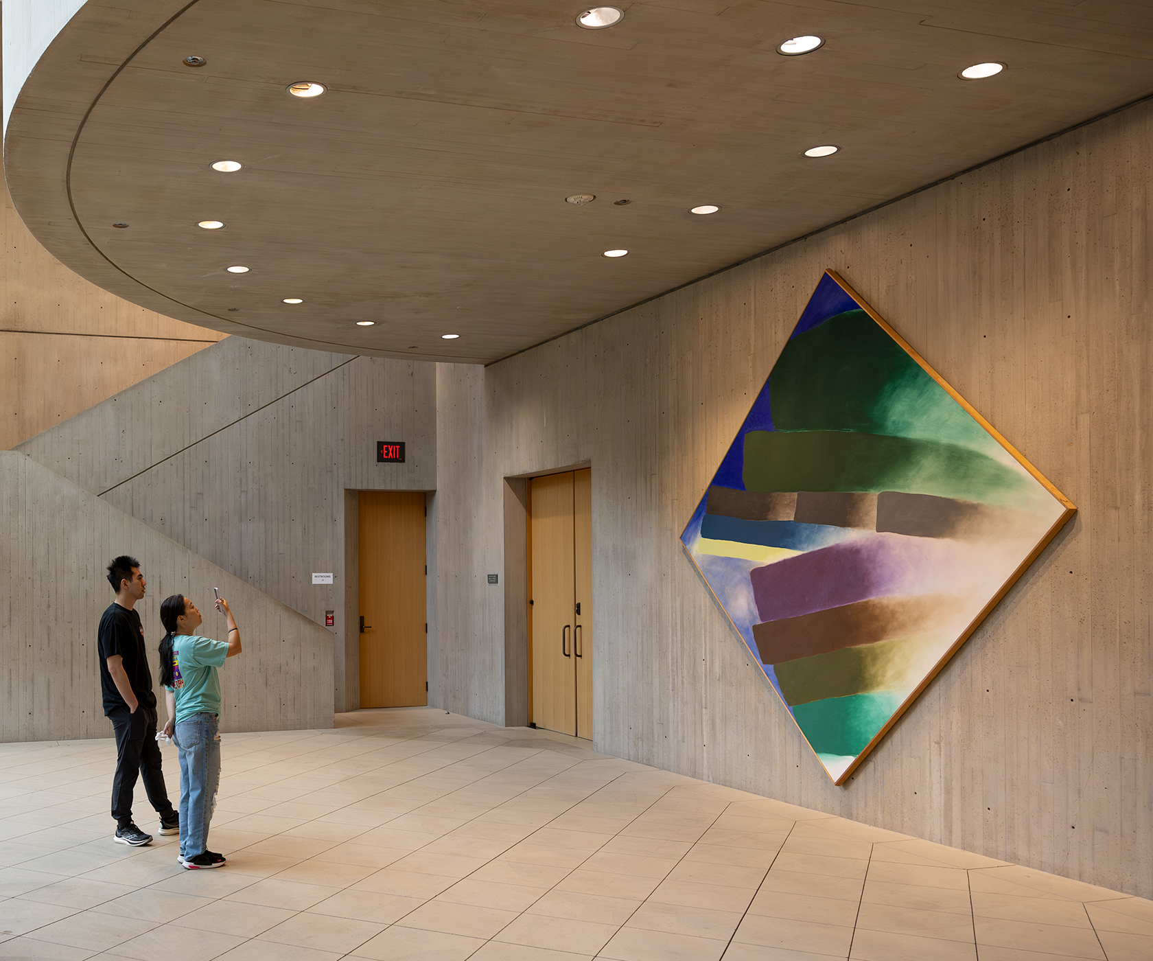 Two people look at a large and colorful diamond-shaped painting in a lobby with concrete surfaces