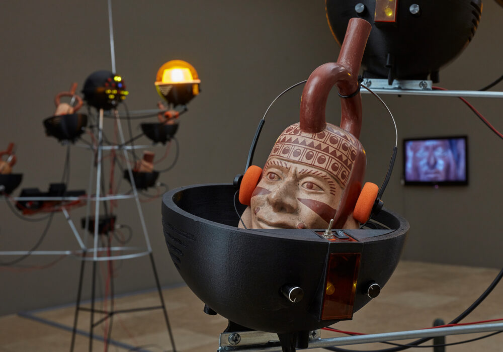A Peruvian ceramic with a human face wears foam-covered headphones and is surrounded by metalwork, lights, and monitors