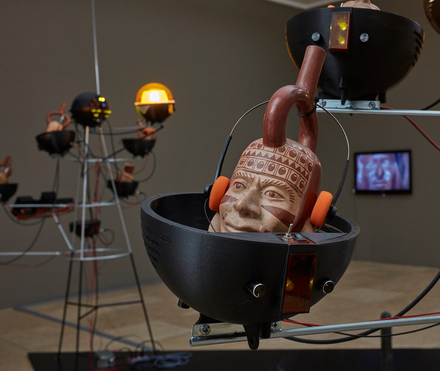 A Peruvian ceramic with a human face wears foam-covered headphones and is surrounded by metalwork, lights, and monitors