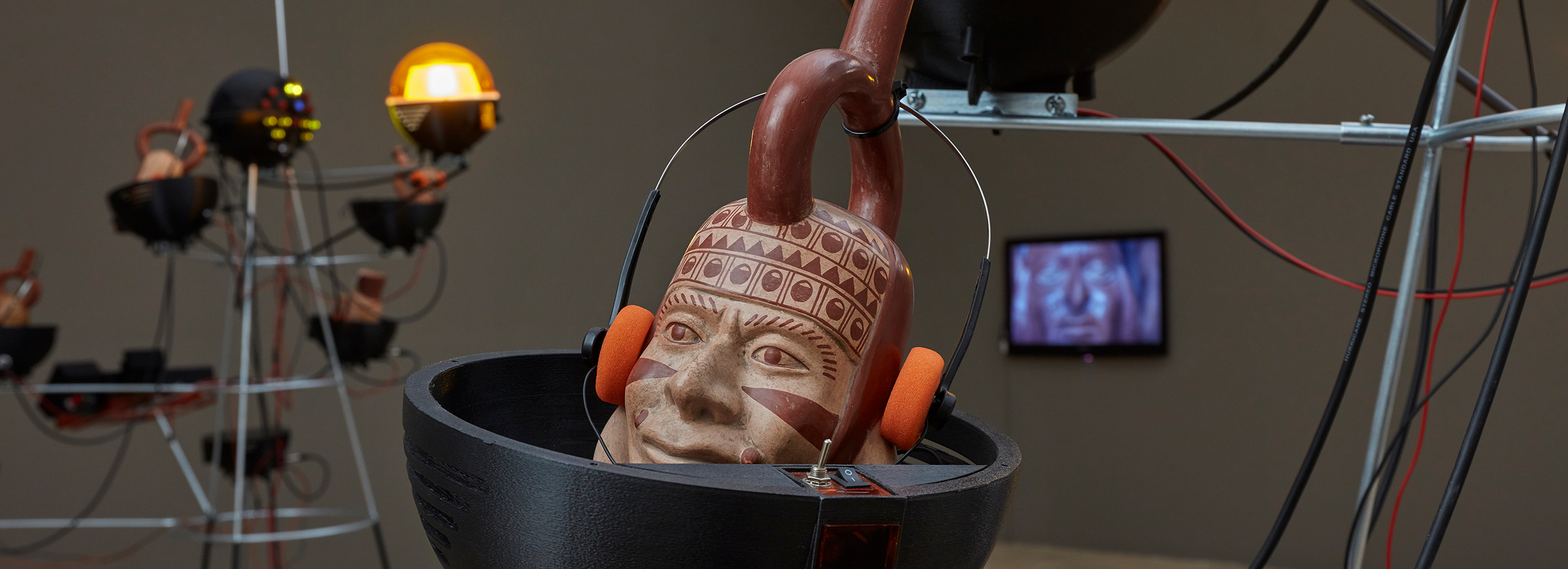 A Peruvian ceramic with a human face wears foam-covered headphones and is surrounded by metalwork, lights, and monitors