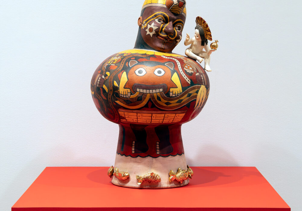 A painted figurative sculpture with a tiny cherub on its shoulder displayed on a red table