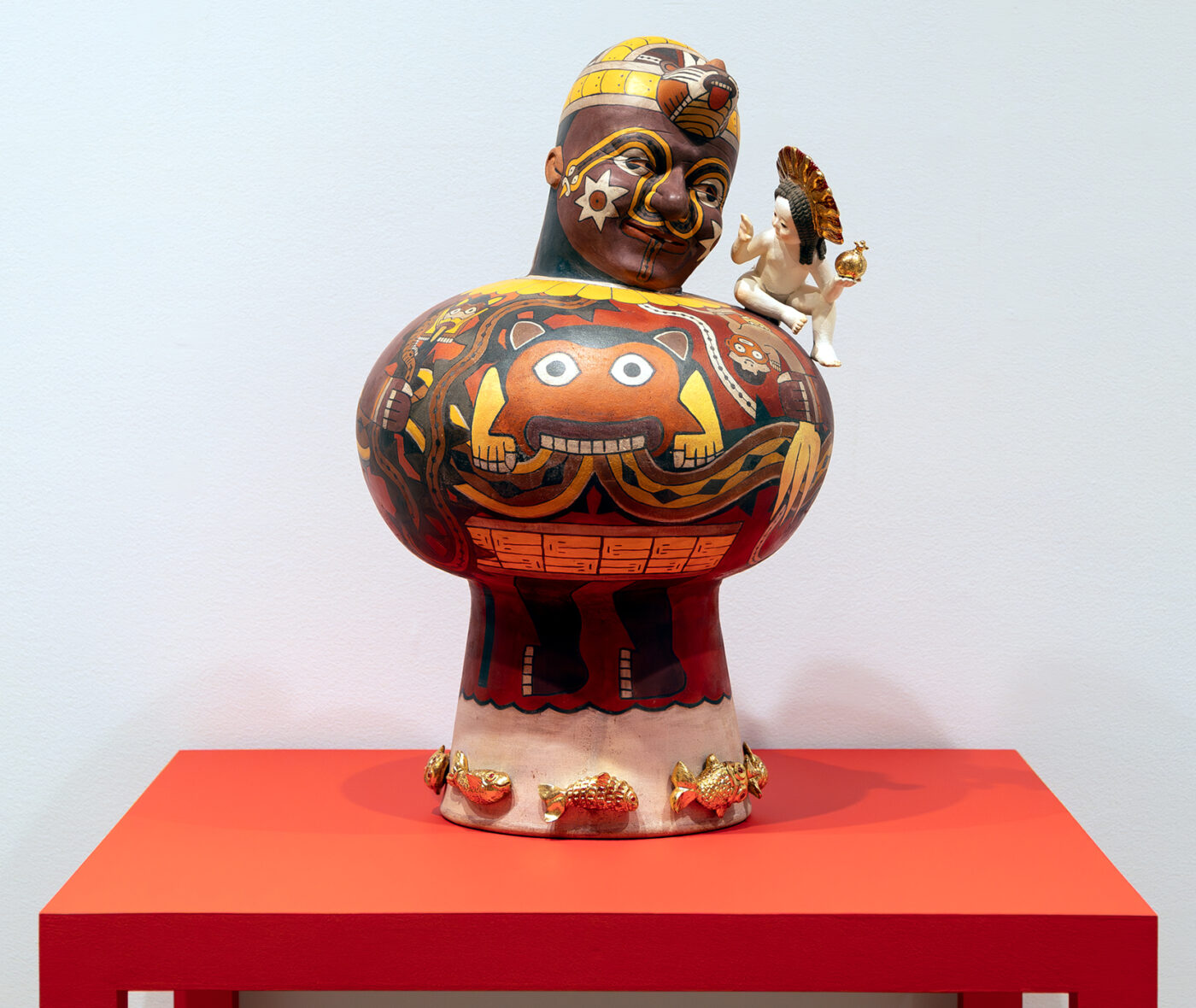 A painted figurative sculpture with a tiny cherub on its shoulder displayed on a red table