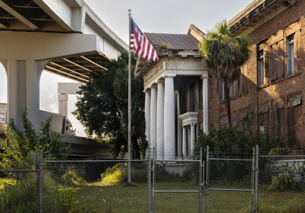Color photograph of an abandoned school building and a flying American flag under an elevated highway