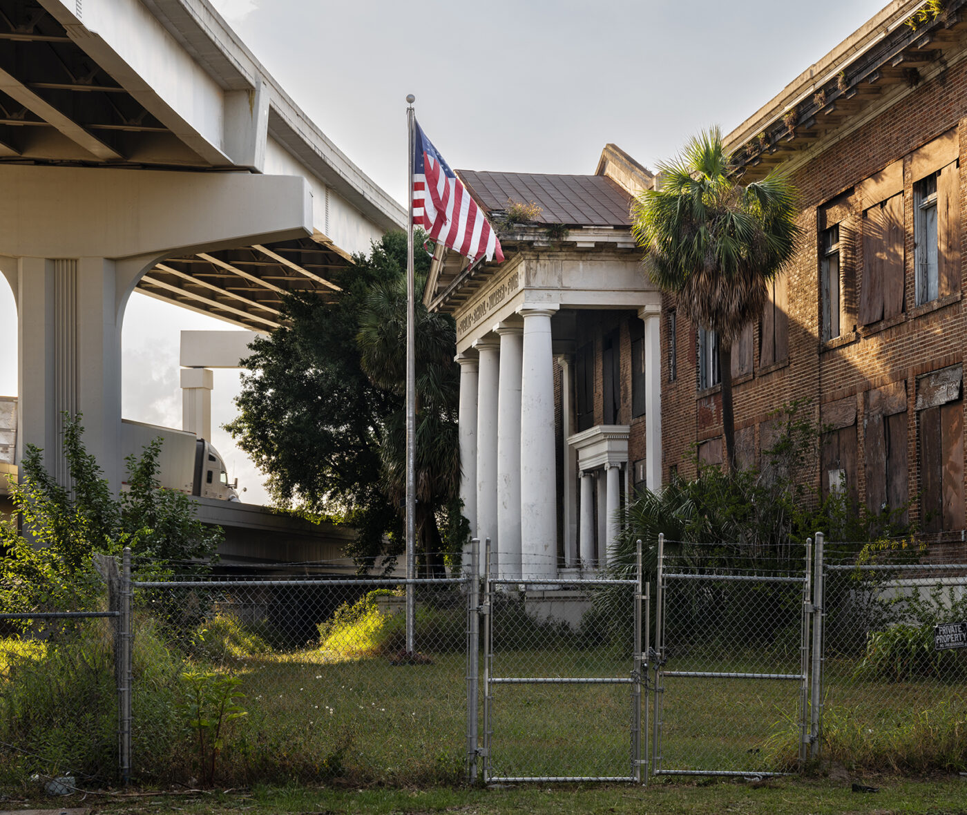 Color photograph of an abandoned school building and a flying American flag under an elevated highway