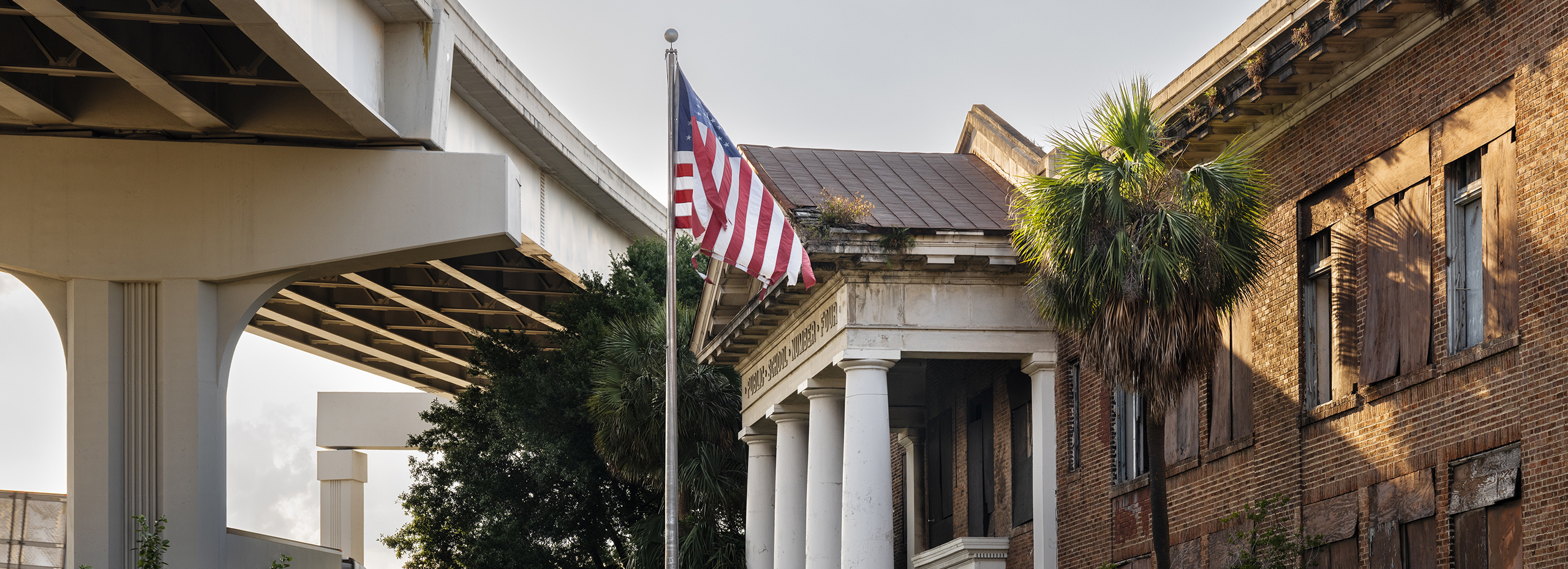 Color photograph of an abandoned school building and a flying American flag under an elevated highway