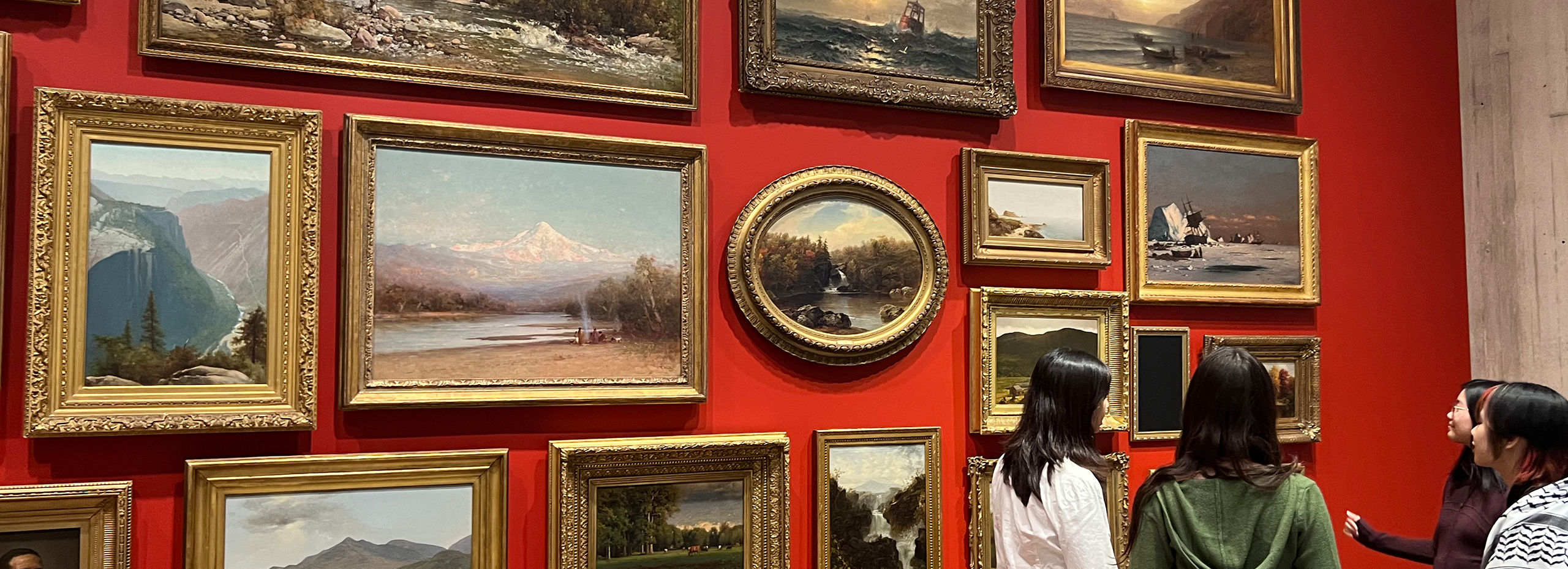 A group of students stand before a red wall filled with gold-framed paintings