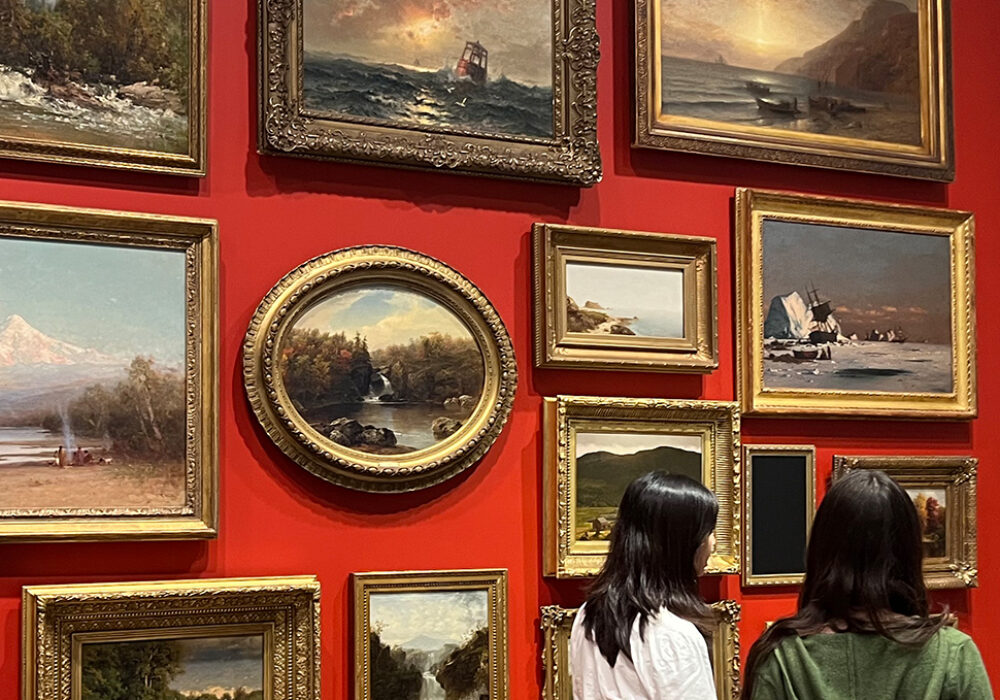 A group of students stand before a red wall filled with gold-framed paintings
