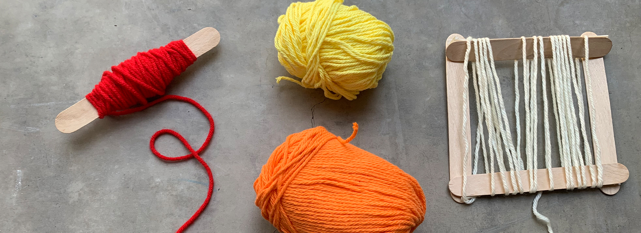 Tools for weaving including red, orange, and yellow yarn, a spindle, and a popsicle-stick loom