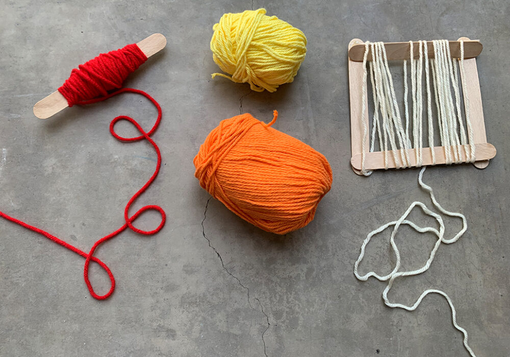 Tools for weaving including red, orange, and yellow yarn, a spindle, and a popsicle-stick loom