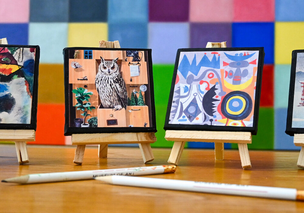 Mini artworks on easels and white pencils on a table in front of a colorful painting