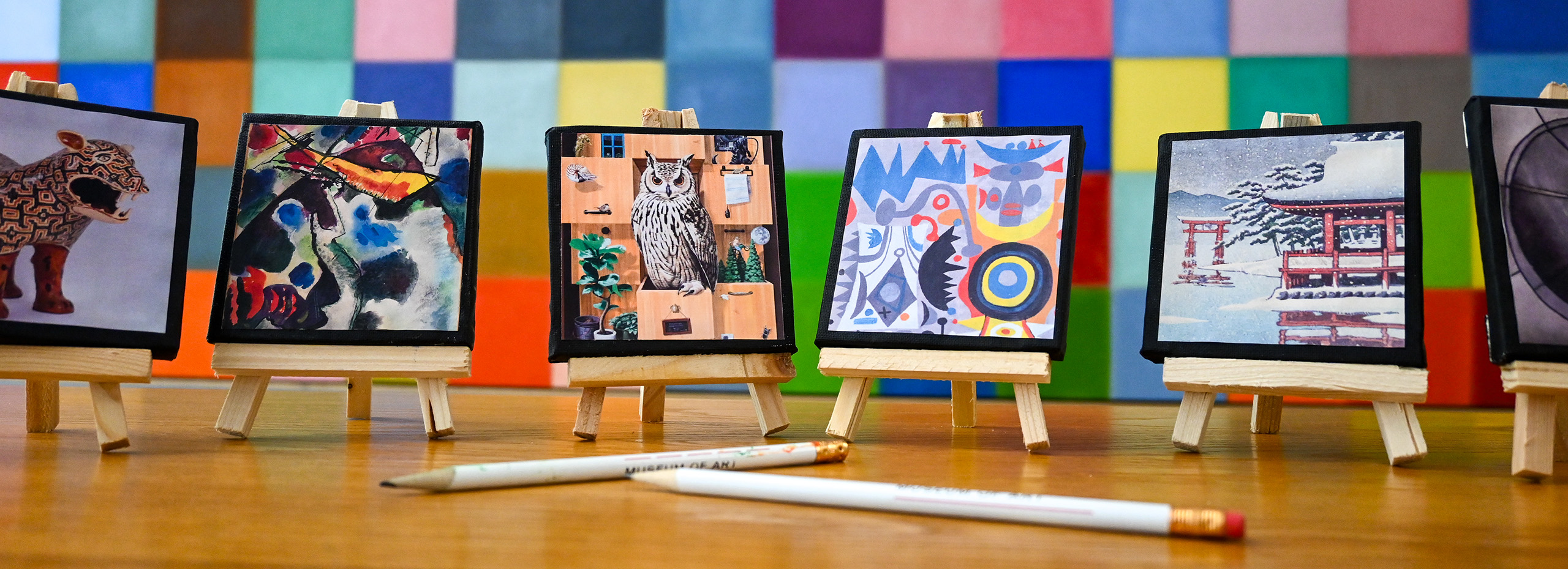 Mini artworks on easels and white pencils on a table in front of a colorful painting