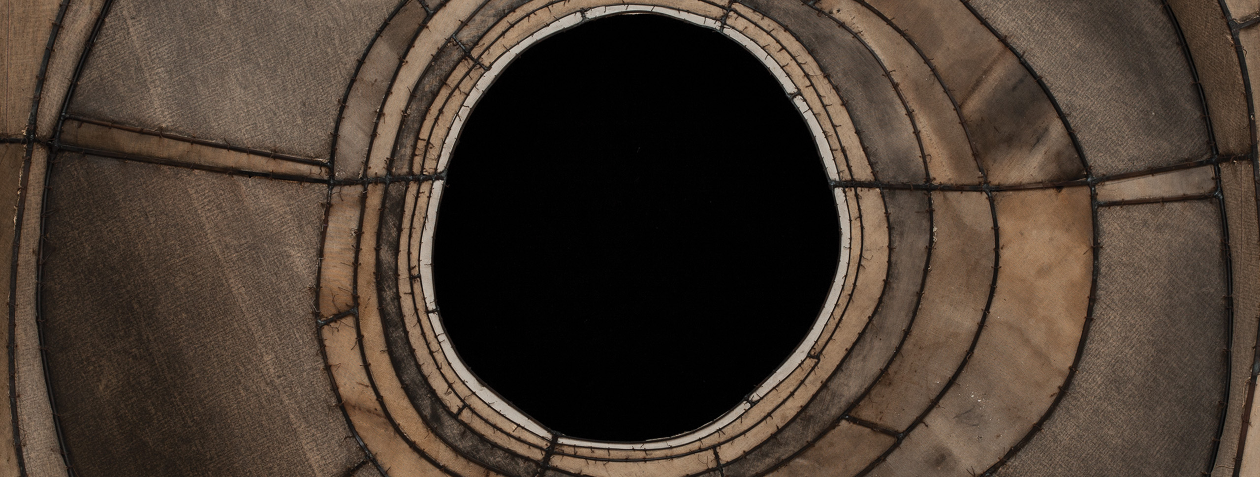 Portion of a hanging sculpture made of stitched brown canvas with a black hole at the center