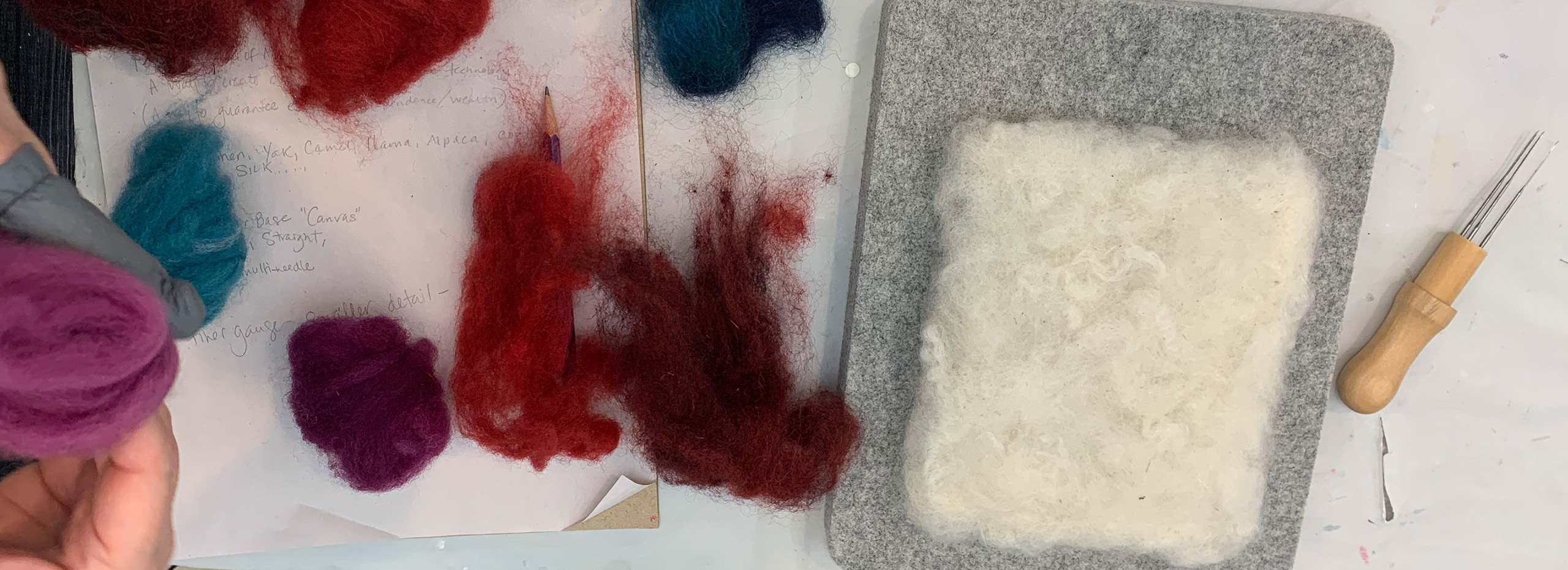 Piles of colorful wool, needle tools, and a felt block for crafting